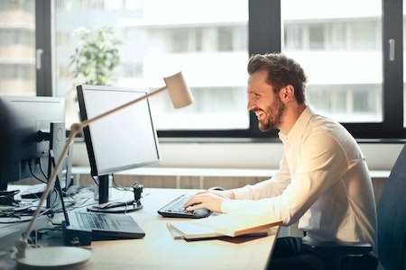 Man in White Dress Shirt Sitting on Black Rolling Chair While Facing Black Computer Set and Smiling - office jobs for college students